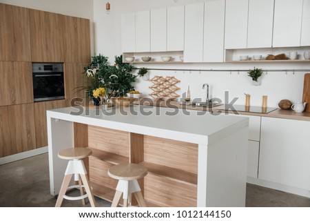 Stylish kitchen in white and brown wood. Style minimalism. Sink, table top, plants, pot, shelf for dishes. Bar stools, table.