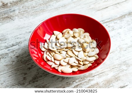 Many pumpkin seeds on a red bowl over a wooden table, background