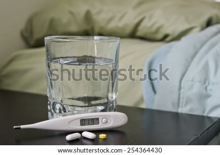 Thermometer showing fever with medicine and cup of water next to bed