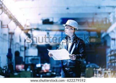 A young successful engineer with a drawing in his hands is standing in the territory of a modern factory. blue toned image