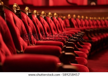 Traditional classically regal ornate rounded wood armed formal plush deep red velvet opera movie theater chairs in curved row with decorative gold molding in fancy carpeted venue