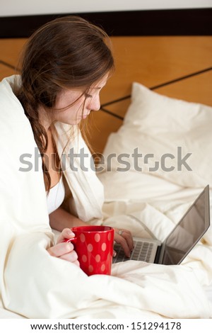 Young lady just woke up sitting in bed checking her laptop computer and having a cup of coffee.