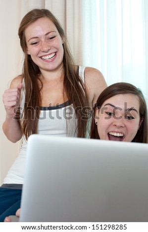 Two young women are sitting, looking at a laptop and laughing out loud.