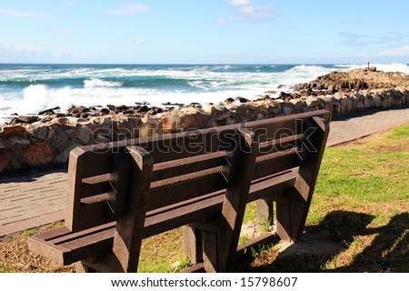 An empty wooden bench next to a pathway that runs next to a rocky shore, on a sunny Winters day.