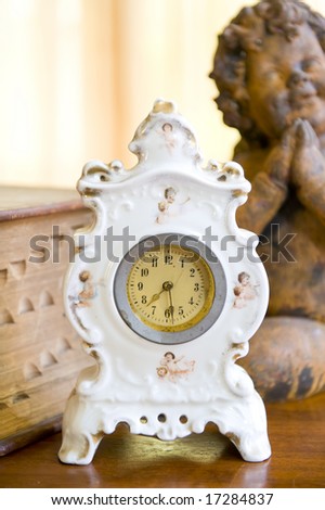 An antique clock on a wooden table in front of an old reference book and brass statuette of an angel.
