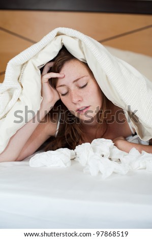 A sick young lady in bed, lying with her head under the blankets and a pile of tissues in front of her.