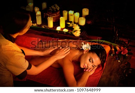 Massage of woman in spa salon. Girl on candles background treats problem nack and sholders. Luxary interior with working hands masseuse. Crop of bare back has relax. Alternative medicine on very dark