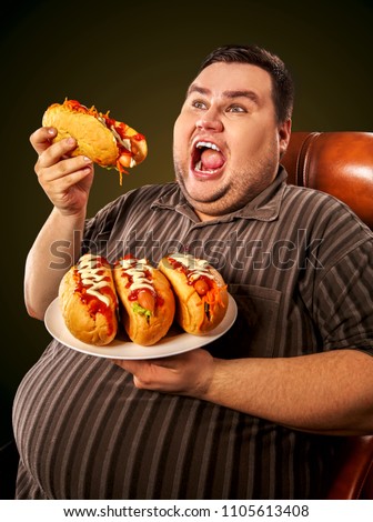 Fat man eating fast food hot dog on plate. Breakfast for overweight person. Junk meal leads to obesity. Person regularly overeats concept on black background. Man after hunger strike.