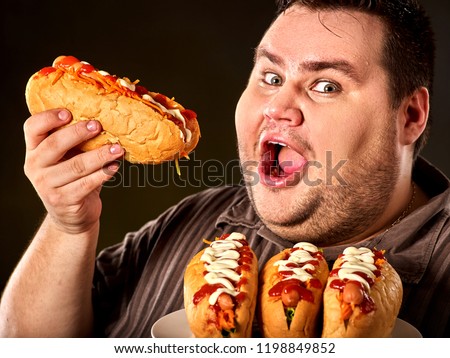 Hot dog contest. Fat man eating fast food hot dog on plate. Breakfast for overweight person. Junk meal leads to obesity. Dangers of extra weight. Person regularly overeats concept on black background.