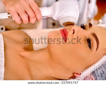 Close up of young woman in hat receiving electric ultrasonic facial massage at beauty salon.