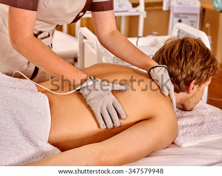 Strong man receiving back electric massage at beauty salon. Beautician wearing electricity gloves.