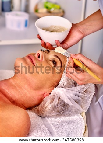 Woman middle-aged in hat take facial and neck clay mask in spa salon.