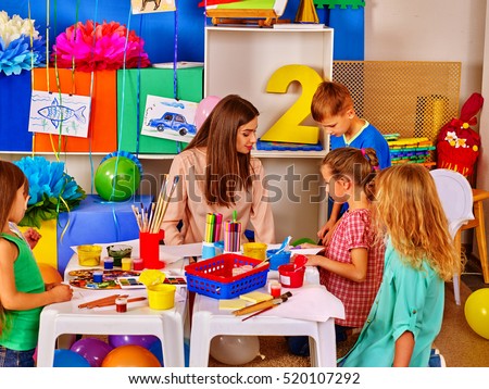 Children with teacher woman painting on paper at table in kindergarten . Children with teacher make overall project.