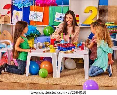 Children painting and drawing in kids club. Art lesson in primary school. Kindergarten teacher who smiling help small students. Kid boy coloring picture on table in class. Balloons on floor.