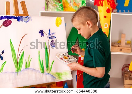 Child painting on easel. Kid boy learn paint by brush in class school. Kindergarten interior on background. Boy is getting ready to become an artist.