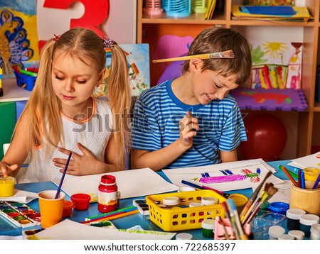 Small students girl and boy painting in art school class. Child drawing by watercolor paints on table in kindergarten. Boy is holding a brush behind his ear.