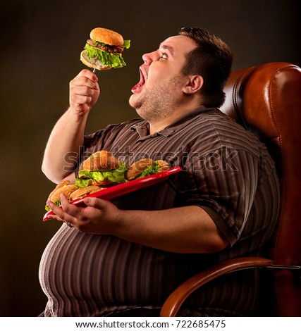 Diet failure of fat man eating fast food hamberger. Happy overweight person with wide-open mouth greedily eating huge hamburger on fork. Hate to diets. Business chairs for fat people concept.