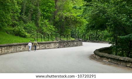 A middle aged couple walk down a curving rural road lined with a stone wall and lush green foliage.