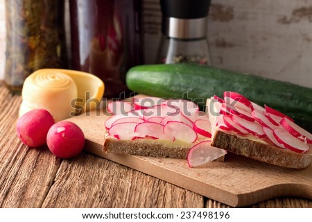 Picture of bread slice with butter and cut radishes. Steamed rolled chease and cucumber is placed near wooden board and jam with preserved herbs are in background.