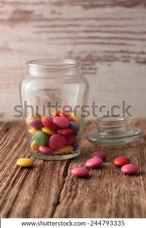 Color chocolate smarties inside opened glass dose and few pills spilled around on old worn wooden board with white wood board in background.