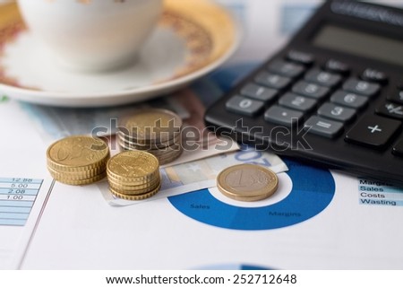Horizontal photo of one euro coin in the middle of pie chart with other coins in stacks and bills under black calculator with coffee cup in background placed on paper sheets.