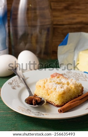 Vertical photo of Fresh apple cake and star anise on spoon placed on green worn wooden table with flour, butter, eggs and empty glass bottle