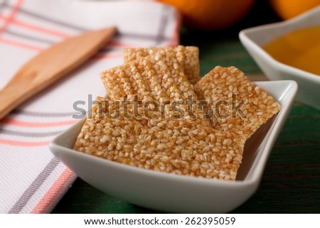 Horizontal photo of Sesame wafers with honey in white bowl, placed on green wooden board and cloth with wooden spoon on left.