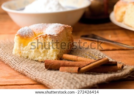 Horizontal photo of single portion of curd fresh cake on jute cloth together with few pieces of cinnamon. Powder sugar in white bowl and spoon in background. All placed on wooden table.