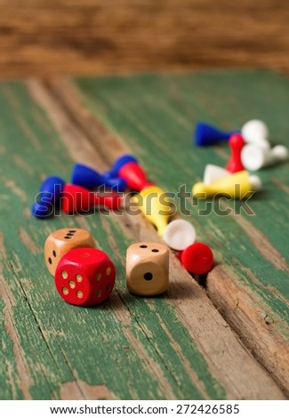 Vertical photo of three dices in front of other color figurines for ludo game. All pieces are placed on old wooden table with worn green color.