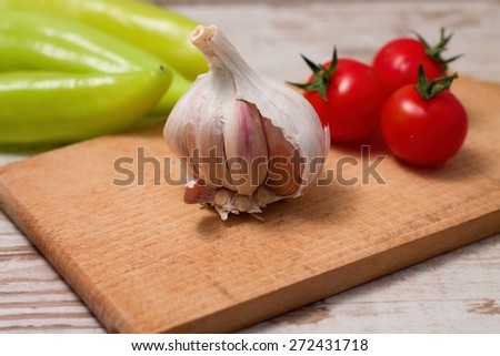 Horizontal photo of single garlic bud and three red tomatoes on chopping board with three green paprica in background. All placed on wooden table with white color.
