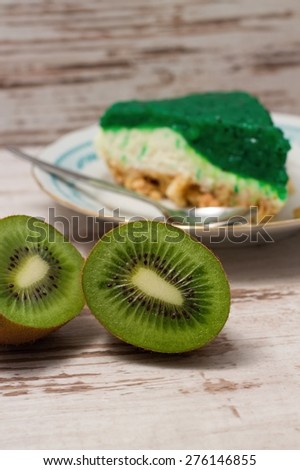 Vertical photo of kiwi fruit cut in half in front of portion of green jelly pie on plate with spoon. All is placed on white table.