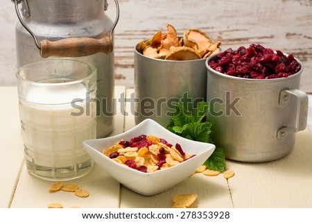 Horizontal photo of scene consists of white square bowl with cornflakes, two cups with dried berries and apples, glass of milk, green herbs and aluminum can. Few cereals are spilled around.