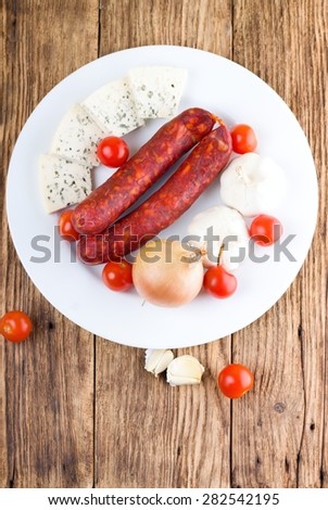 Vertical photo of upper view on white plate with sausages, onions, garlics, tomatoes and cheese with few pieces of vegetable around. All is placed on wooden board table.