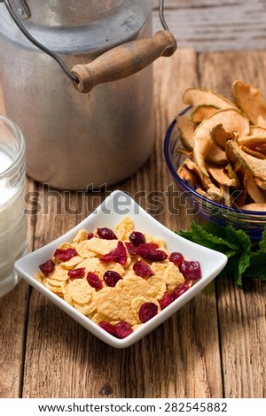 Vertical photo with upper view on square white ceramic bowl full of cornflakes with cranberries in. Glass of milk, old can and another glass bowl with dried apples around. All is on wooden board table