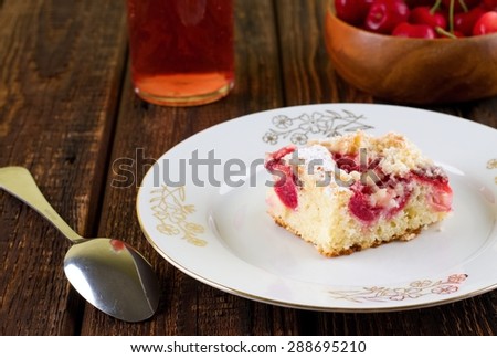 Horizontal photo of white plate with gold motif with single portion of cherry pie on. Around is spoon, bowl with other fruit and red lemonade. All is placed on wooden table.