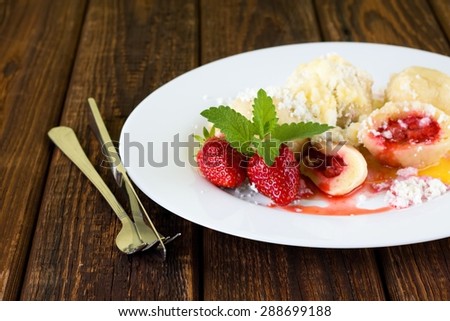 Horizontal photo with white plate full of fruit sweet dumplings sprinkled by sugar and curd with two whole strawberries and herb leaves on side. Knife and fork are on left.