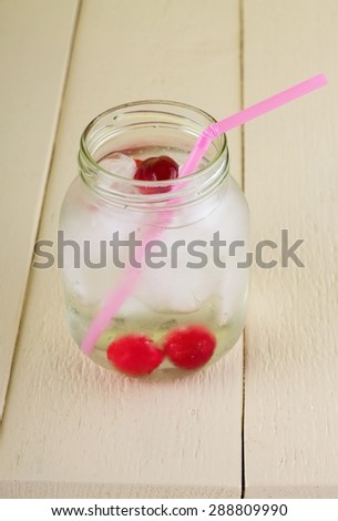 Vertical photo of cold fresh drink from cherry fruit which is inside with ice cubes. Pink straw is too in. All is placed on wooden table board with white color.