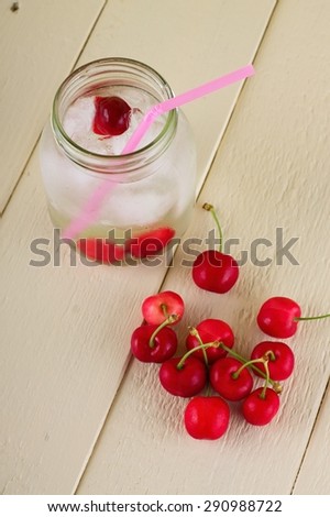Vertical photo of small heap of cherries in front of glass jar full of cold drink, ice cubes and few berries with pink straw. All is placed on white wooden board.
