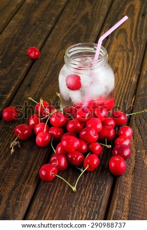 Vertical photo of cold cool drink in a glass jar. Few cherries are in a bottle and heap of them is around. Pink jar is inside. Background is old worn wooden board.