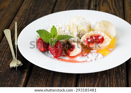 Horizontal photo with fruit dumplings with curd and sugar. Two strawberries with herb leaves are near the edge. Knife and fork are on right and all is placed on wooden table with dark color.