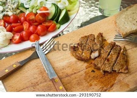 Horizontal photo of pork steak cut in stripes on chopping boar. Knife and fork are next to meat plus bread and several vegetable as tomatoes, cucumber and pepper are around.