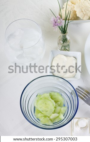 Vertical photo with top view on blue glass bowl with cucumber salad. Fork, glass with ice cold drink, salt and flower plus few pieces of vegetable are around on white cloth.