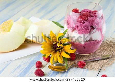 Vertical photo of glass full of two kinds of ice cream in three layers and with raspberries on top which are too around. Glass is on jute cloth with spoon and galia melon in back on blue white board.