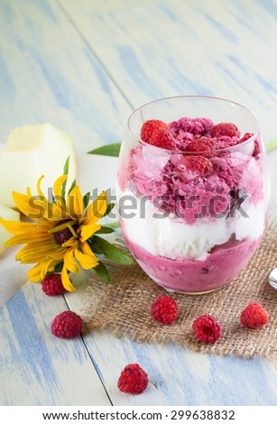 Vertical photo of few raspberries placed around glass with two kinds of ice cream on jute burlap cloth. Yellow flower and galia melon are around on light blue wooden board.