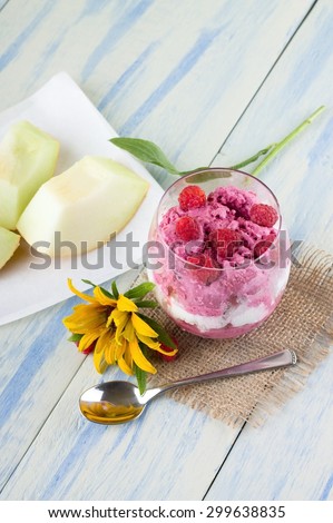 Vertical photo of few raspberries placed around glass with two kinds of ice cream on jute burlap cloth. Yellow flower, spoon and galie melon are around on light blue wooden board.