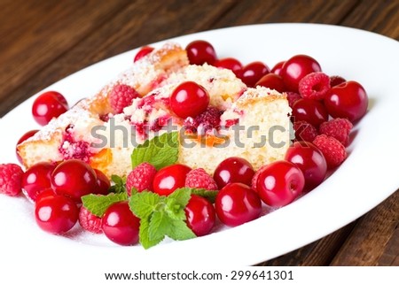 Horizontal photo of single portion fresh fruit cake on white plate with cherries and raspberries and piece of melissa herb together on plate. All is placed on wooden table board.