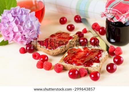 Horizontal photo of two toasts with butter and homemade marmalade among cherries and raspberries. Hydrangea, jar with jam, knife and towel are around on wooden board.