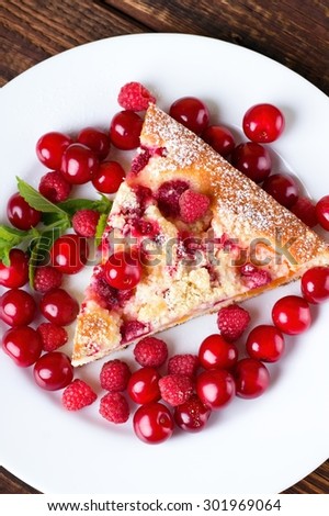 Vertical photo with top view on white plate with single portion of fruit pie / cake and several cherries and raspberries around. Piece of melissa herb is on left. All is on old wooden table.
