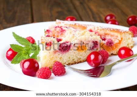 Horizontal photo of single portion of fruit pie with cherries and peaches placed on white plate together with raspberries and other cherries. Green herb and fork are there too.