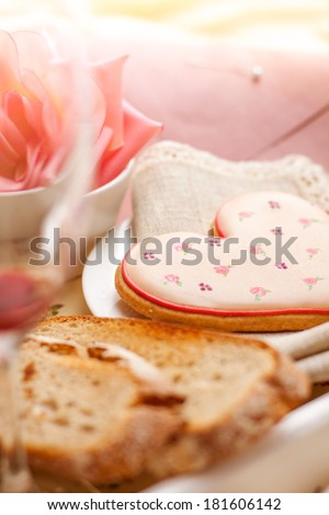 Breakfast in bed detail with heart shaped cookie, fresh whole wheat toast and a rose, with pink envelope in the background. Biscuit decorated in flower pattern with royal icing.